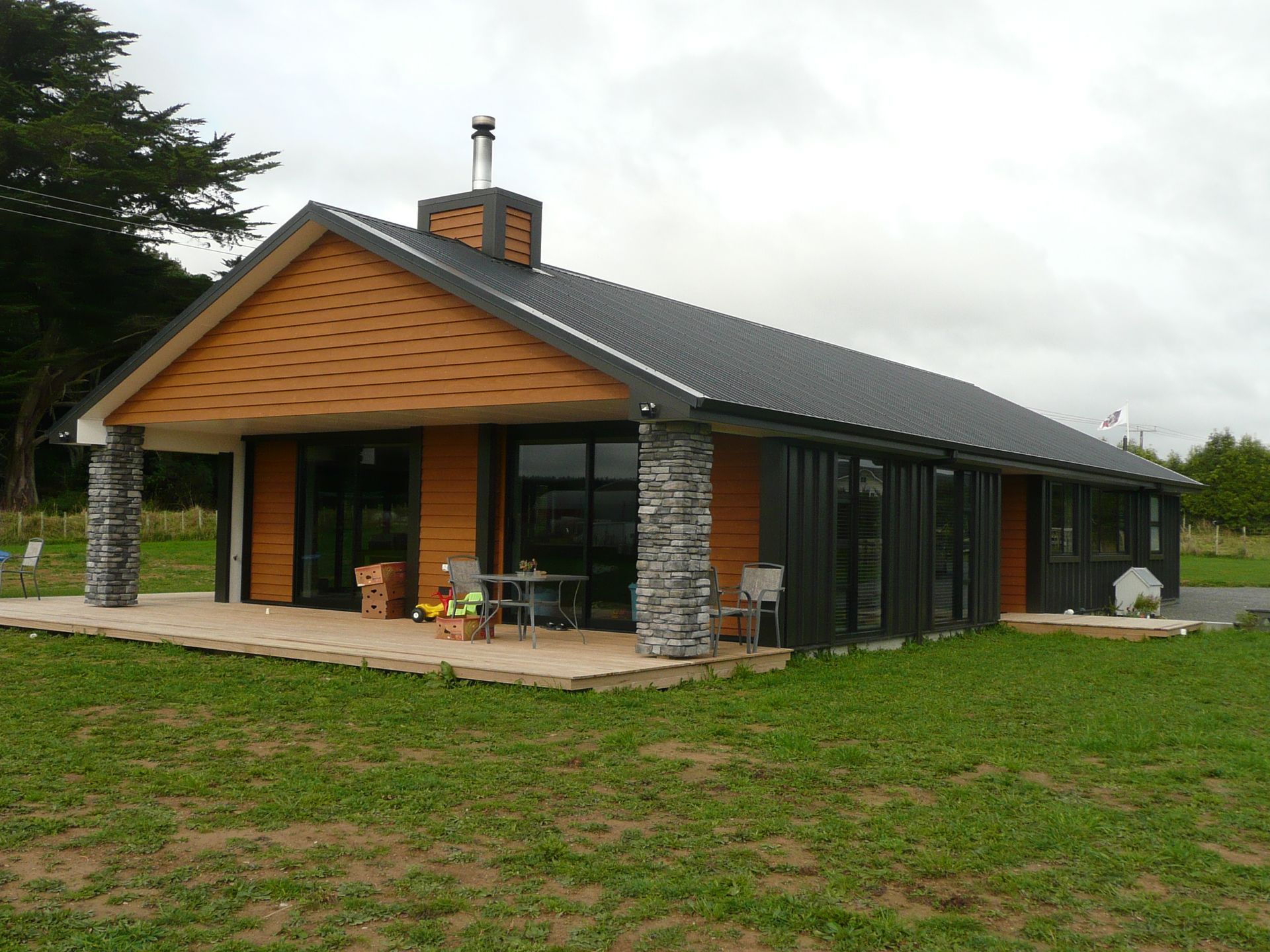 Modern house with gray roof, wooden facade, stone columns, and large windows on a grassy lawn.