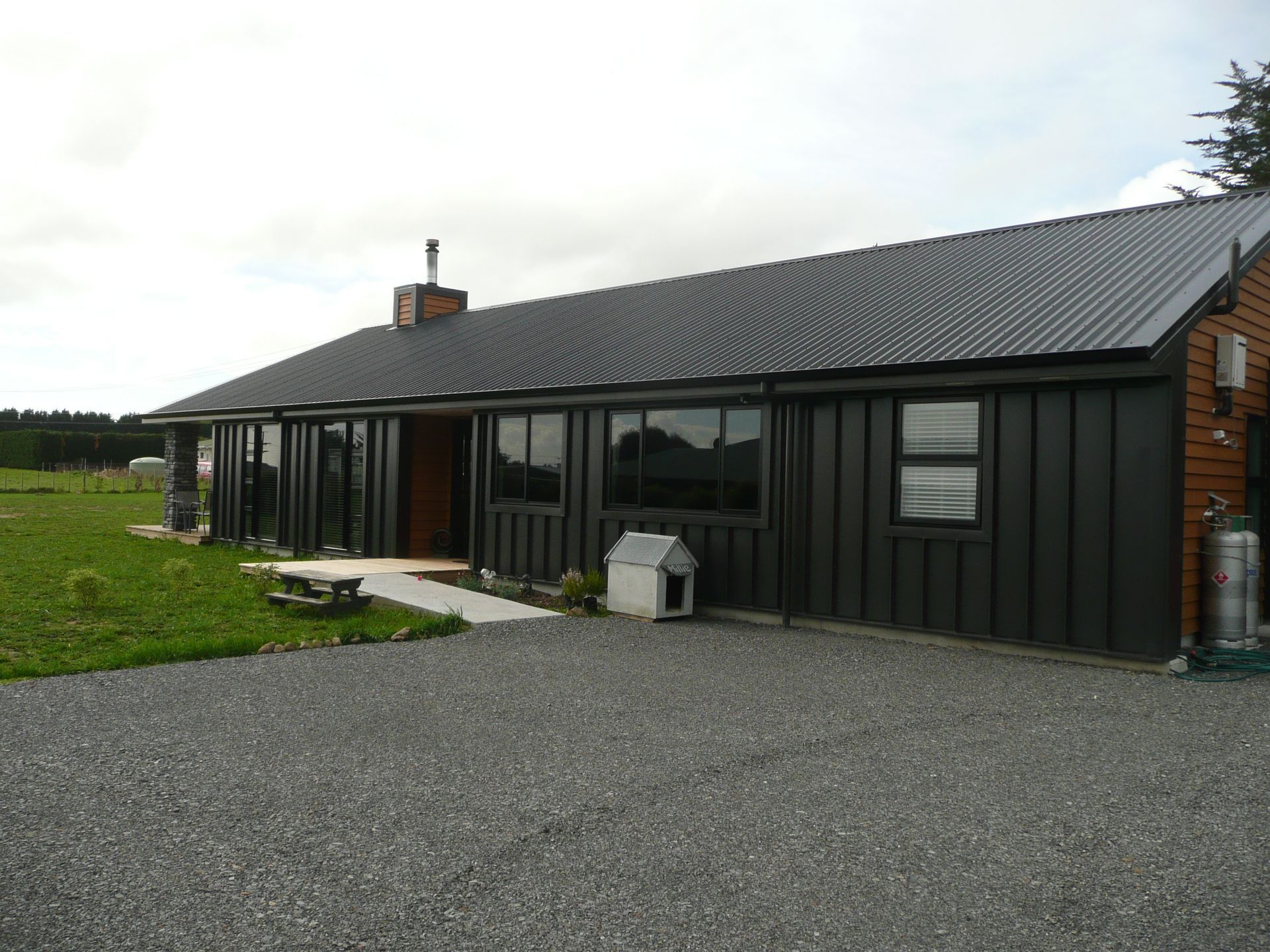 Dark gray house with corrugated metal roof, gravel driveway, and a dog house.