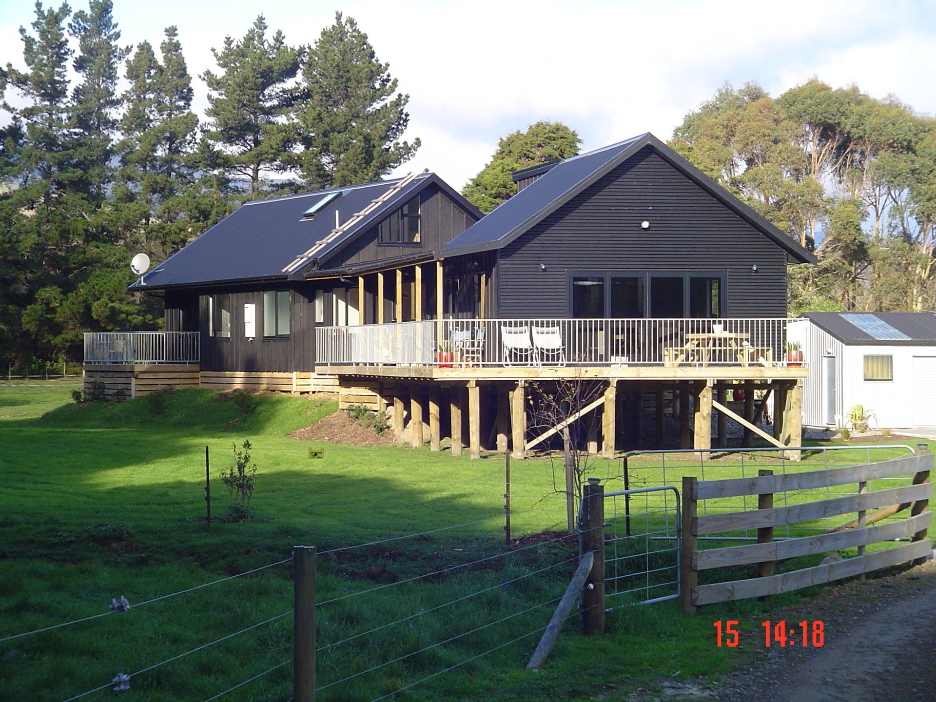 Black house with deck, supported by wooden posts, in a grassy field with a fence.