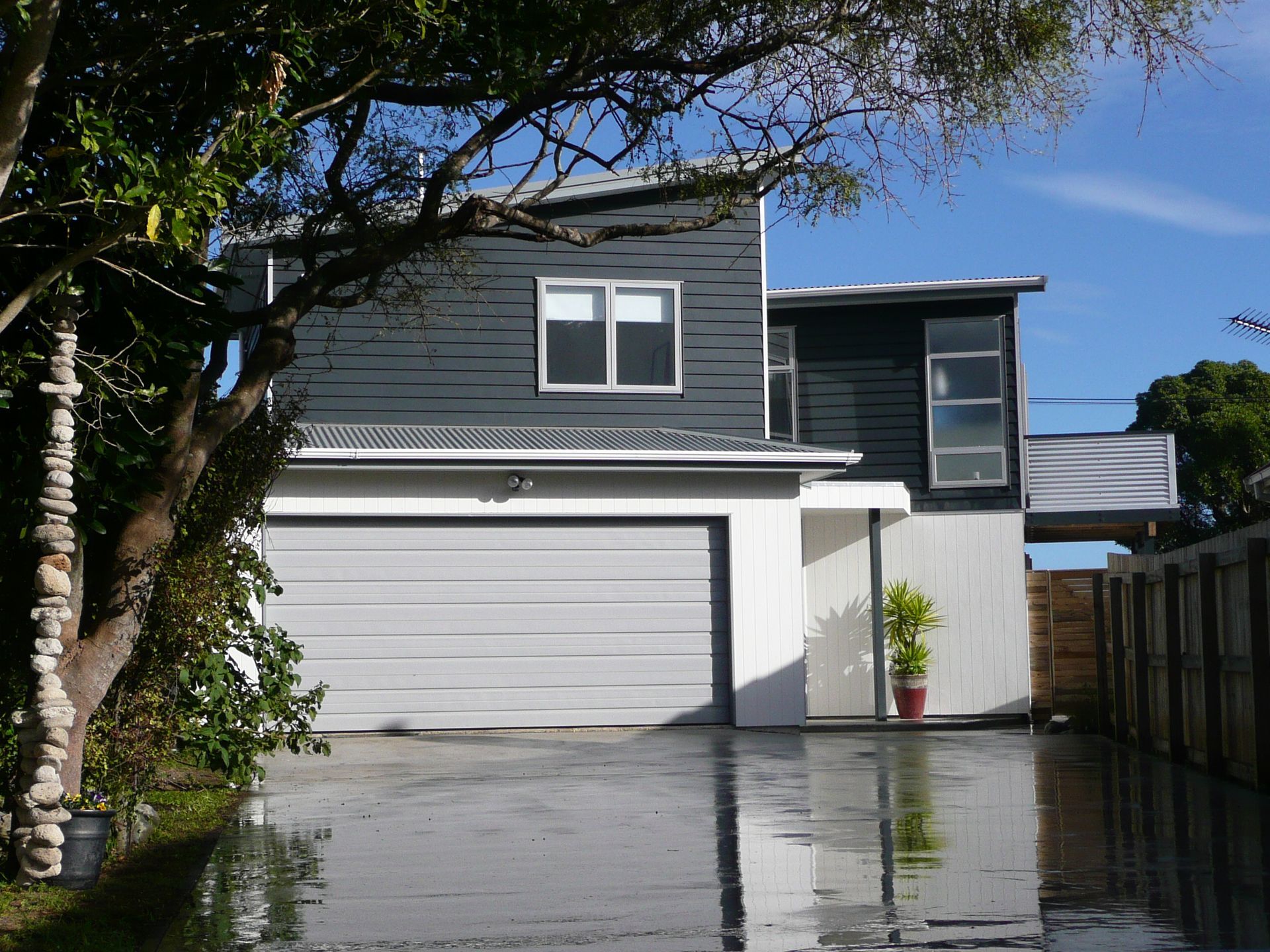 Two-story modern home with a gray garage door, dark siding, and a small tree next to it.