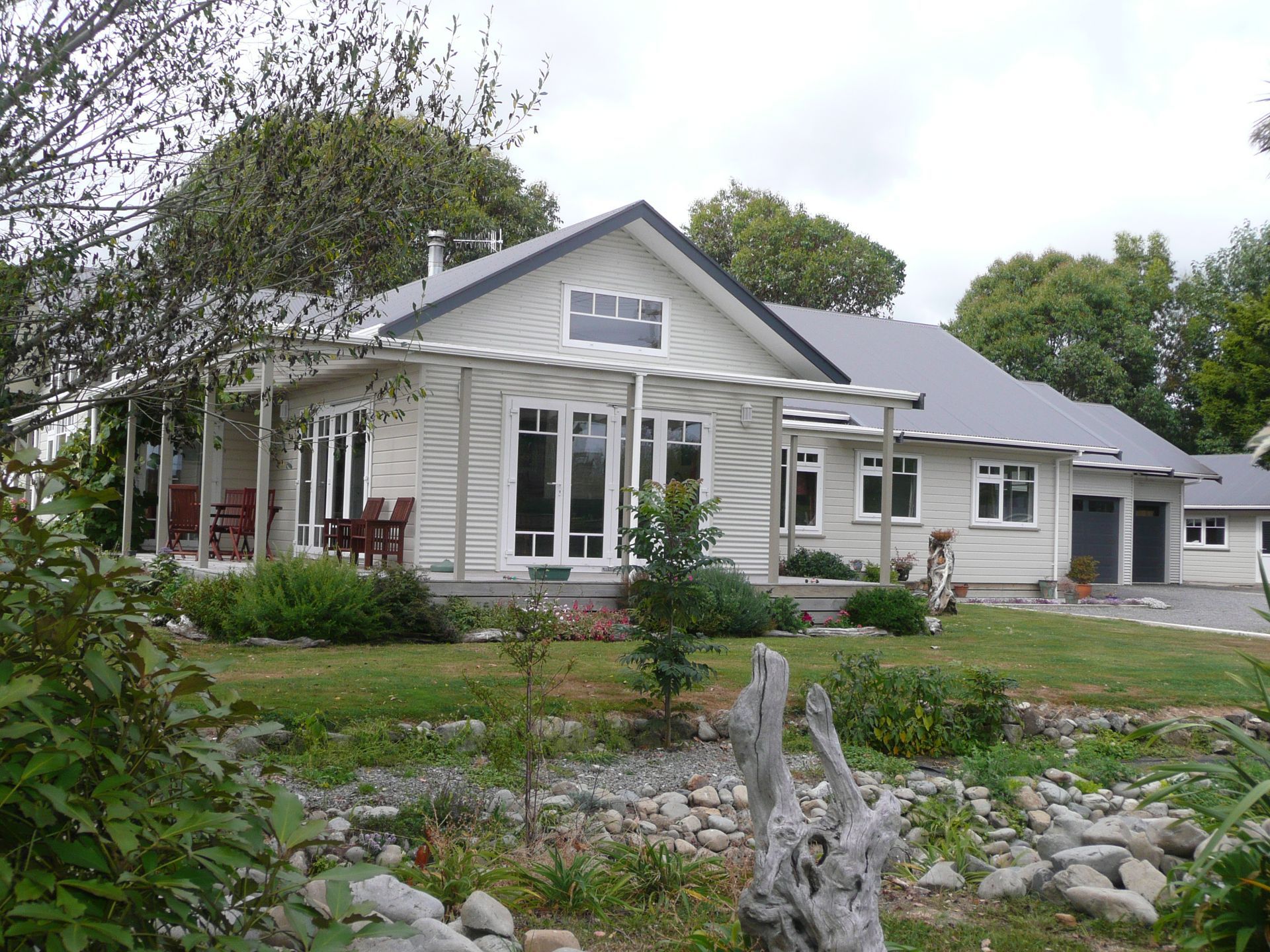 Light grey house with porch, windows, and garage set in a green yard with plants and a cloudy sky.