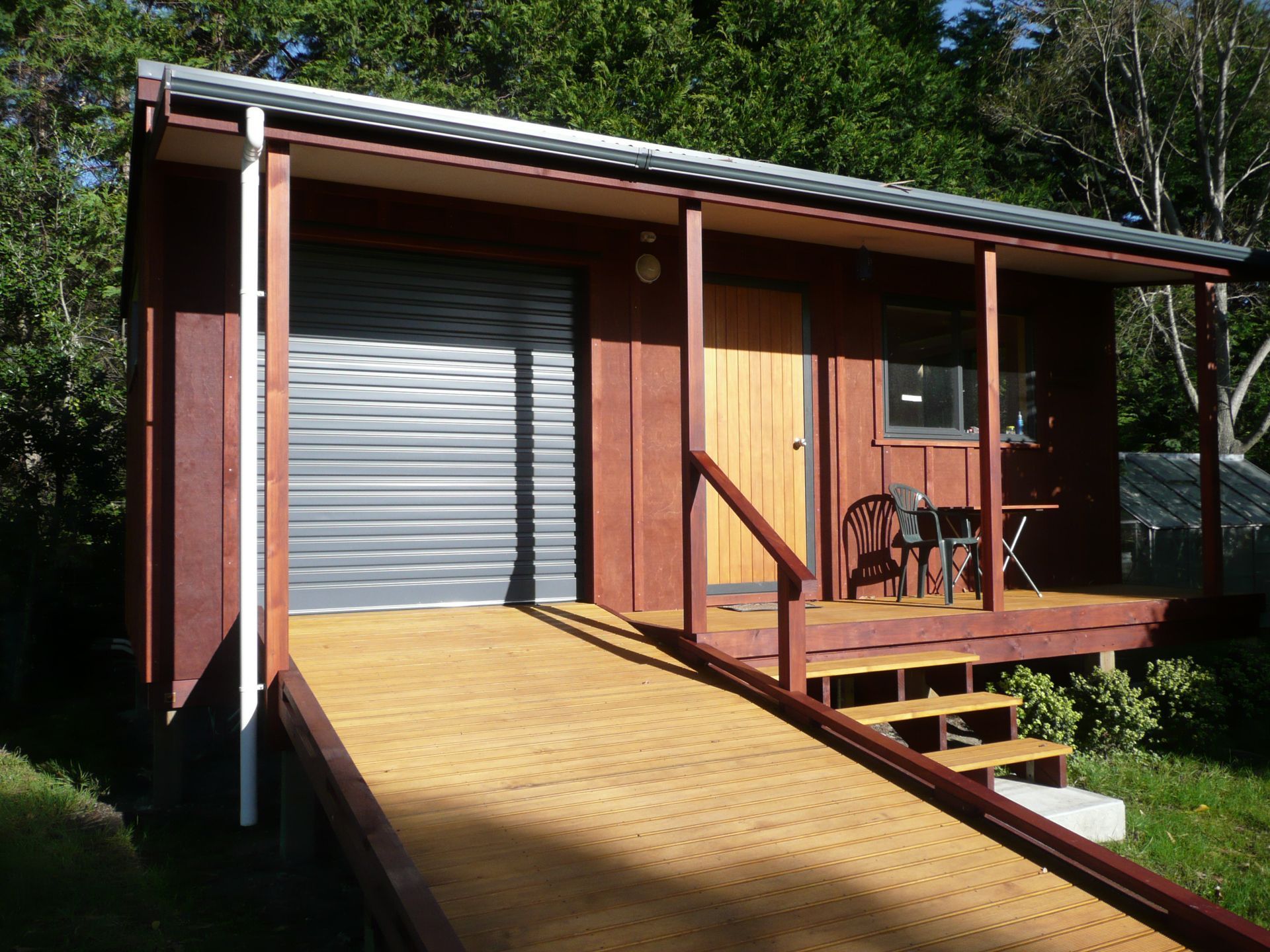 Small brown cabin with ramp, deck, and garage door. Wooden door, porch, and steps visible.