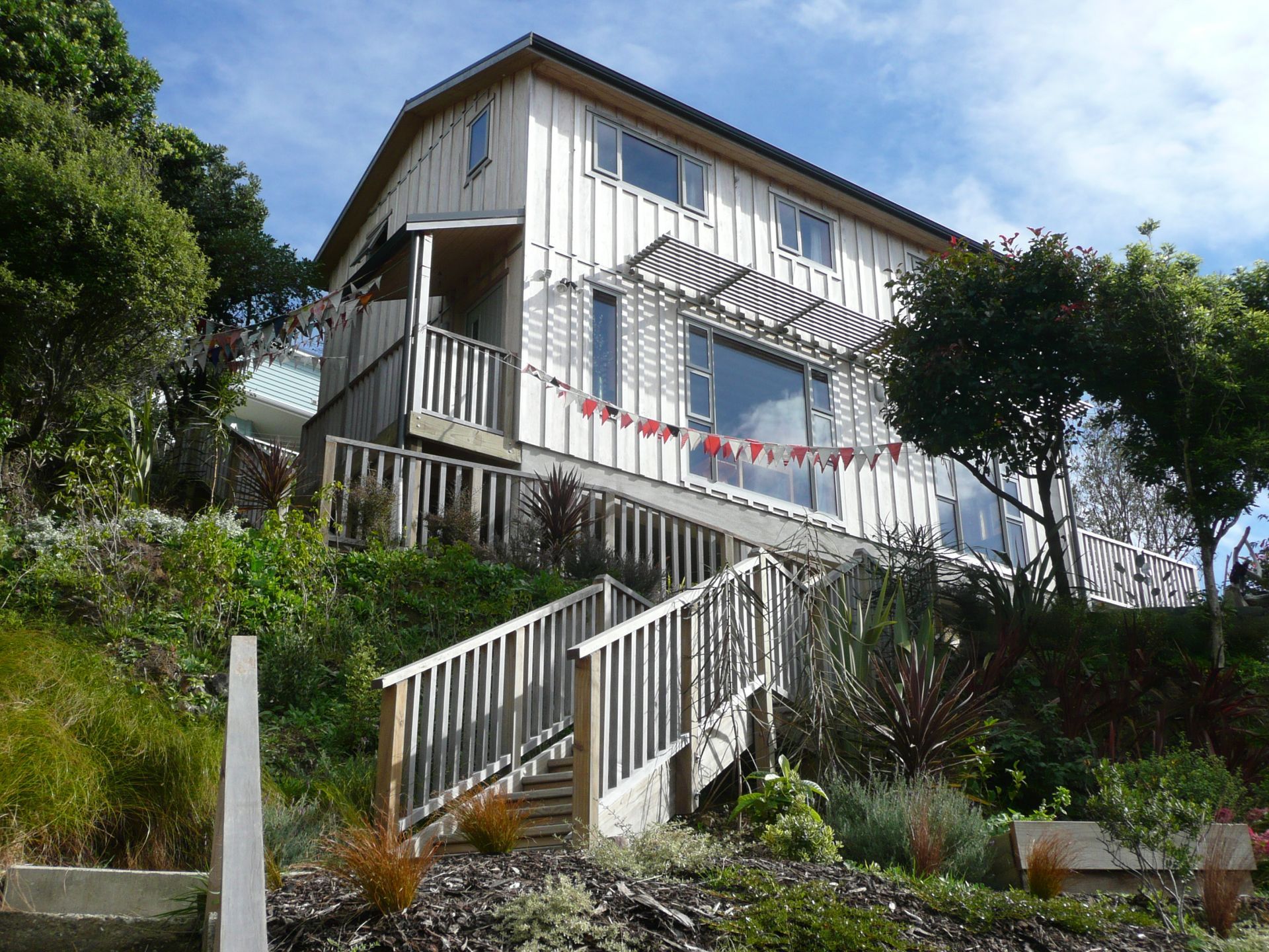 Two-story wooden house on a hillside, with steps leading up to a porch and large windows.