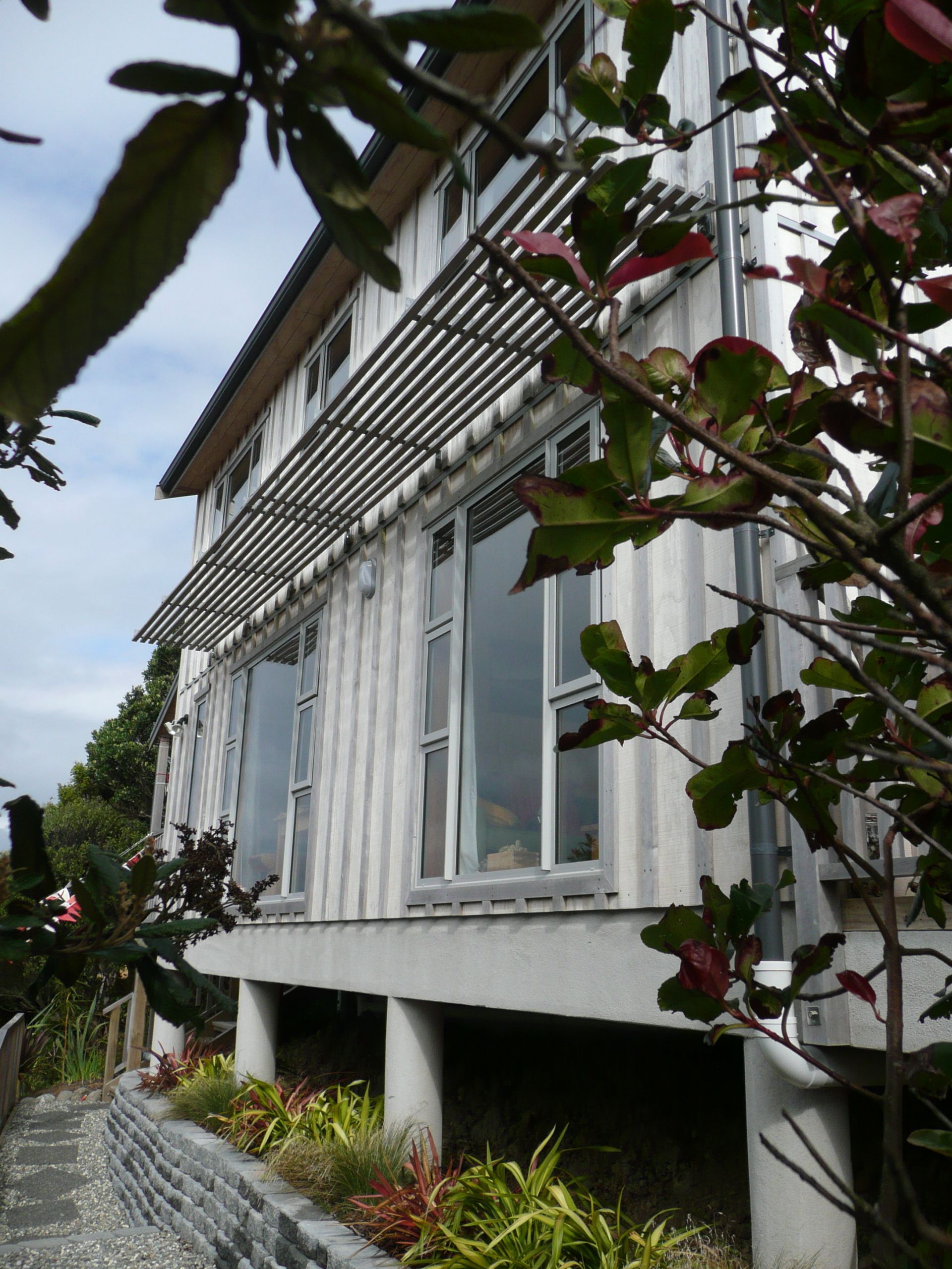 White building with large windows and a slatted awning, viewed from a garden with lush greenery.