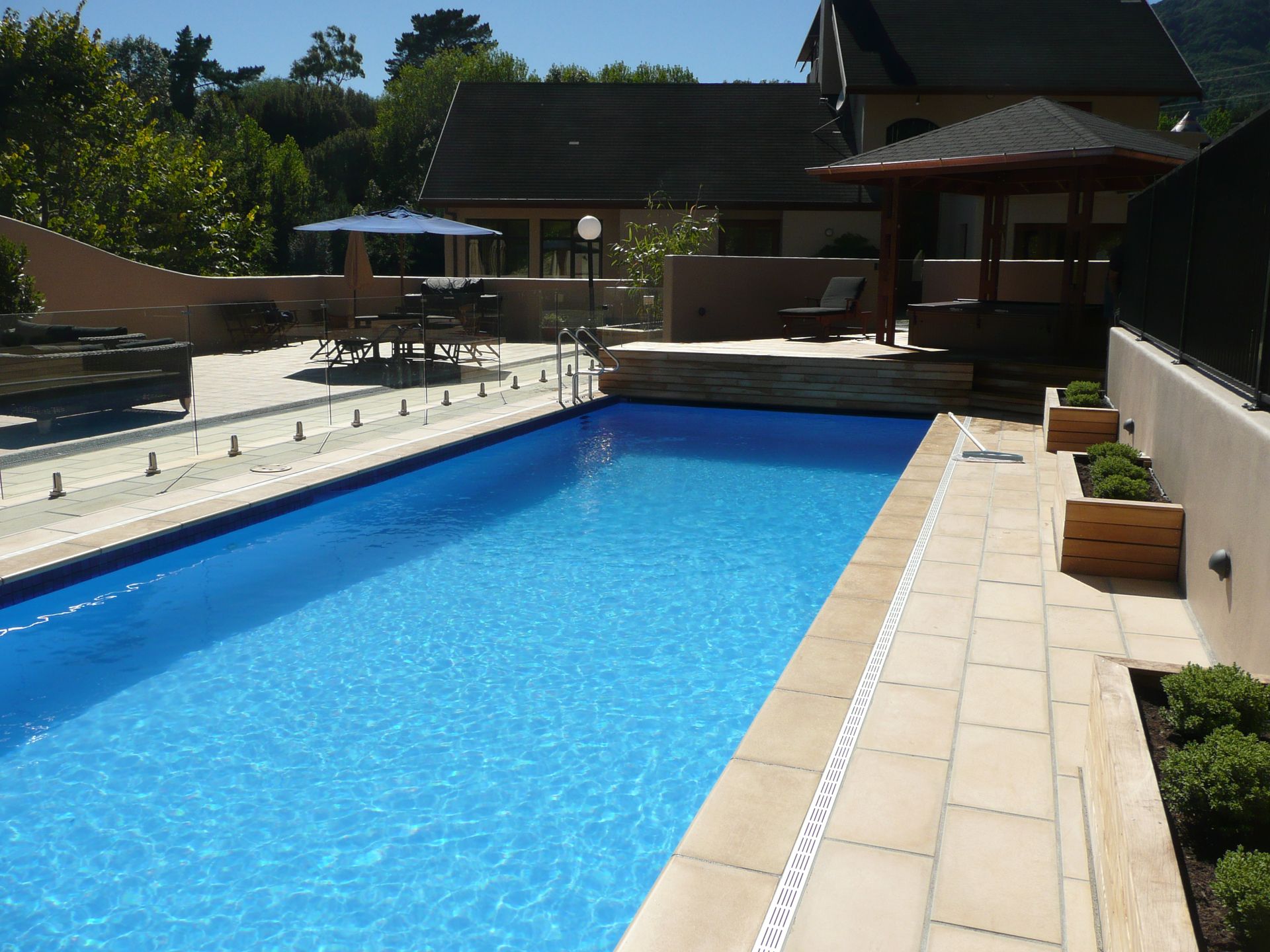 A rectangular swimming pool surrounded by beige stone and a garden, under a sunny sky.
