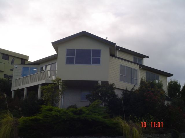 Two-story house with a balcony and large windows, surrounded by green bushes under a cloudy sky.