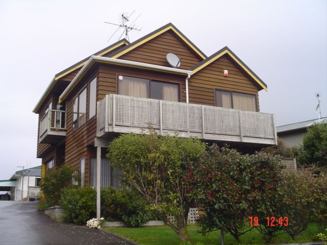 Two-story wooden house with a balcony, shrubs, and cloudy sky.