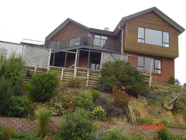 Two-story house with brick base and wooden siding, on a hillside with garden and retaining walls.