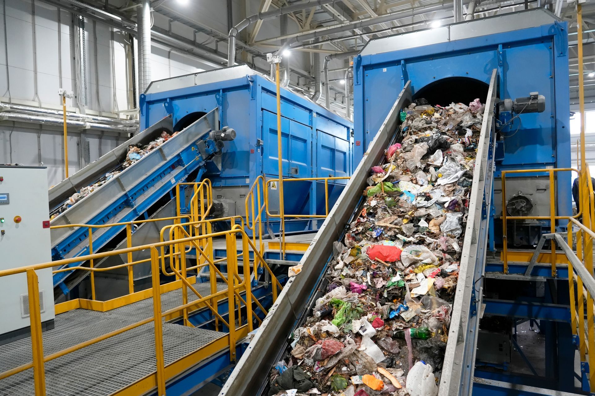 Conveyor belts carrying unsorted waste toward blue processing machines inside a waste management facility.