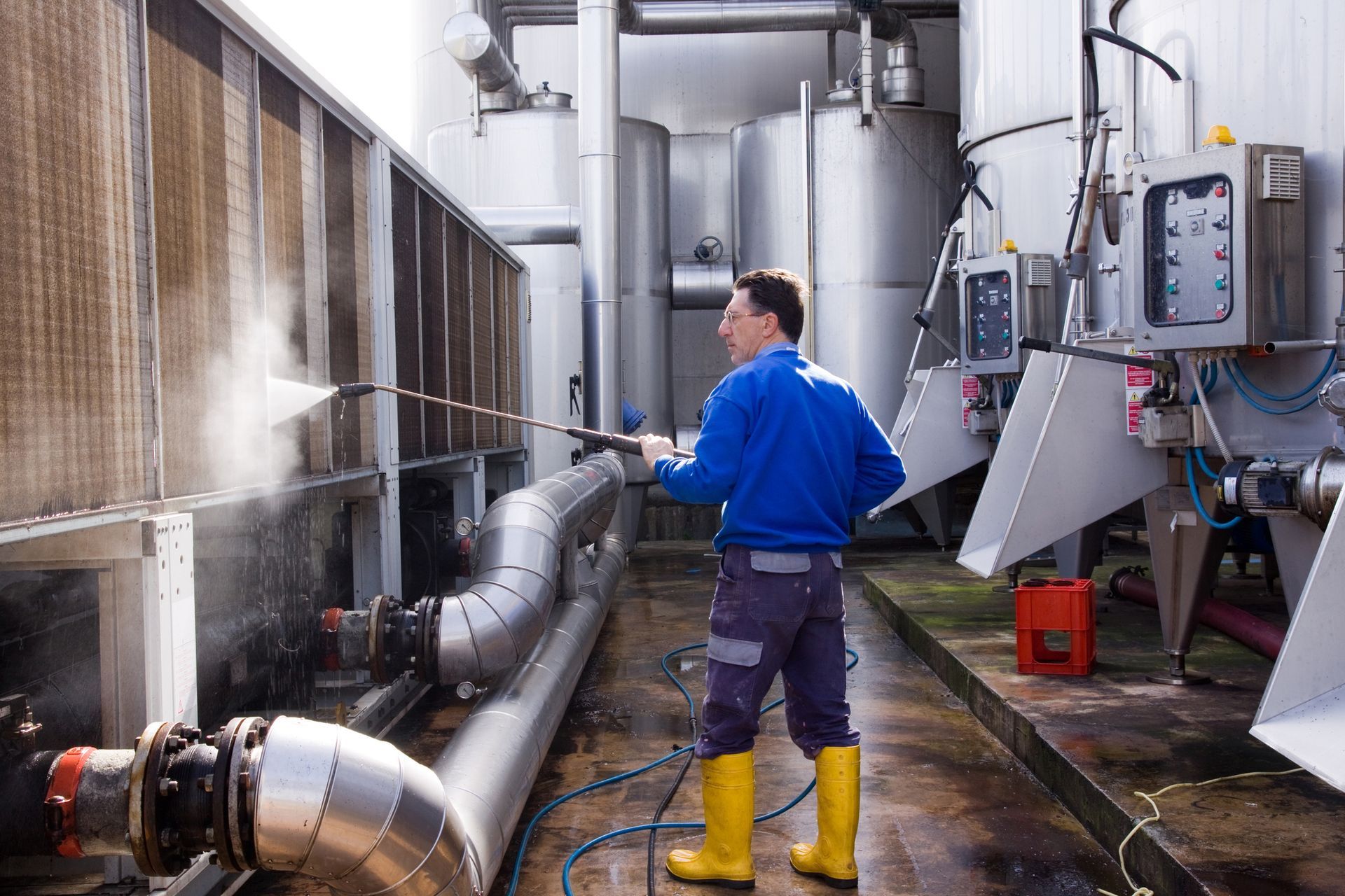 A worker in blue shirt and yellow boots power washes a metal structure in a factory setting.