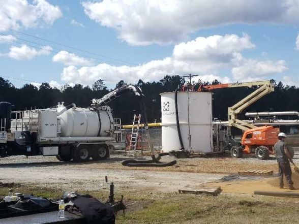 Vacuum truck servicing a large white tank at an outdoor industrial site, with an orange lift, and a worker.