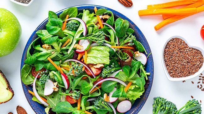 A salad in a blue bowl surrounded by fruits and vegetables on a table.