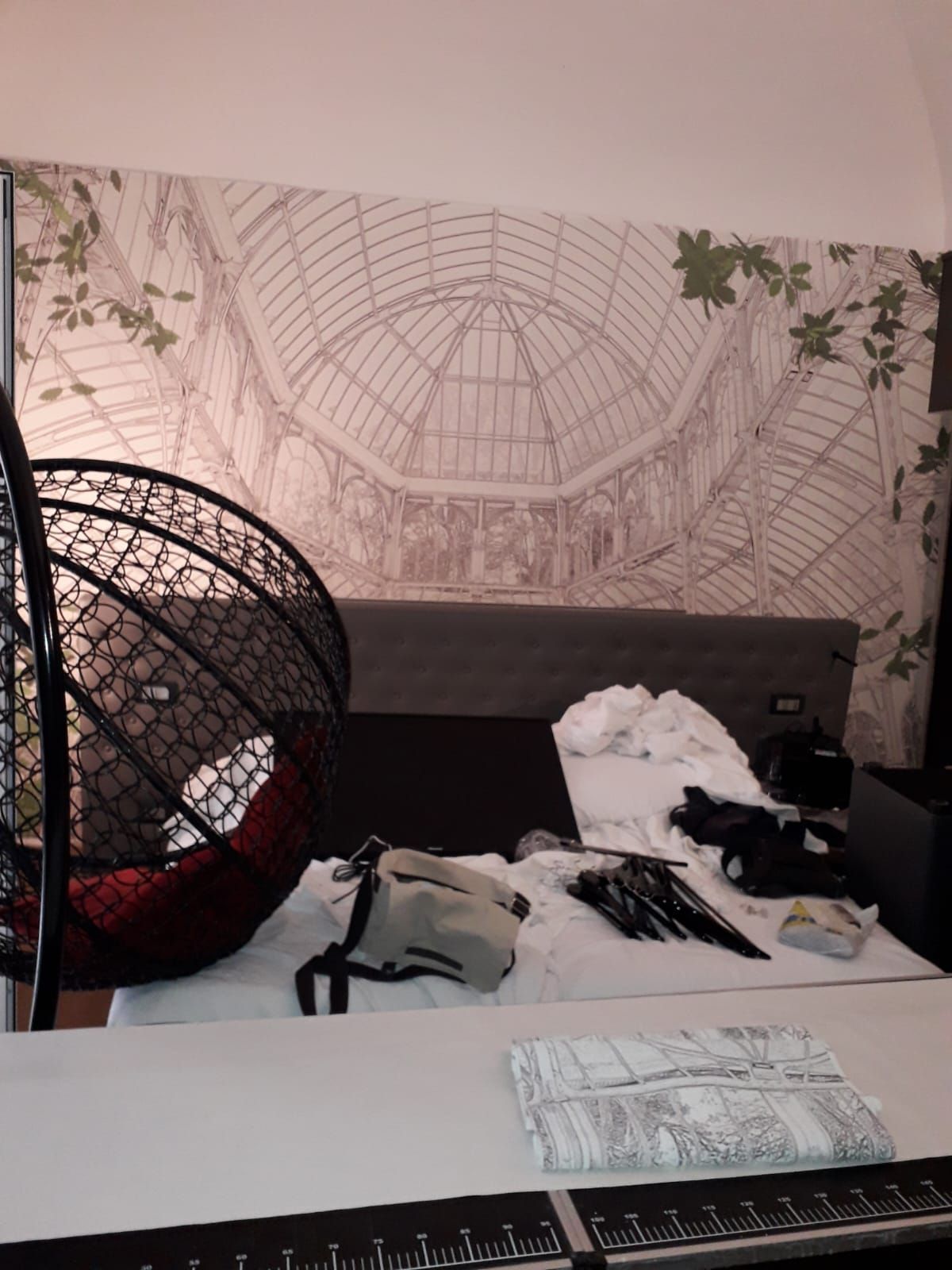 Una camera da letto con una sedia sospesa, un letto con un murale botanico e oggetti su un tavolo bianco.