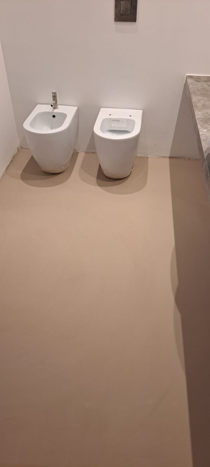 Due water bianchi contro una parete bianca, in un bagno. Pavimento beige, un piccolo elemento decorativo.