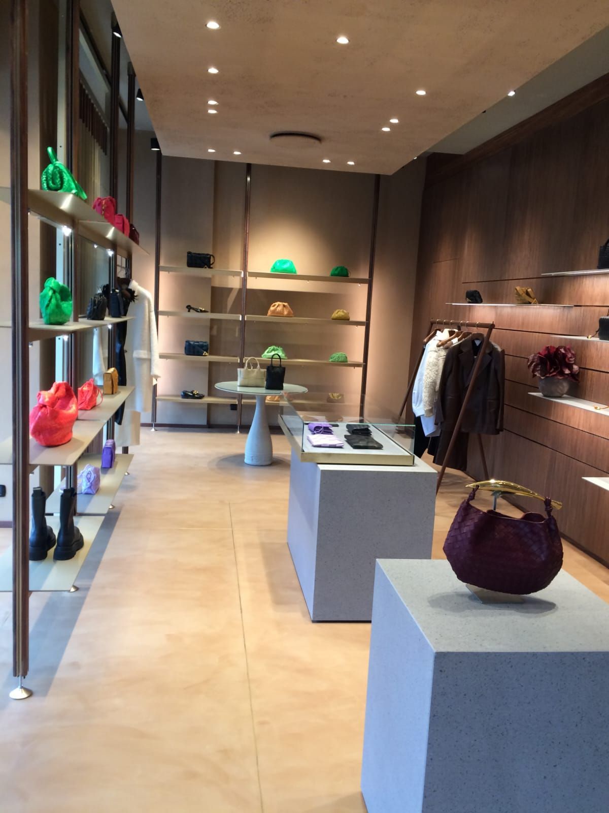 Interno di un negozio di lusso con scaffali che espongono borse e vestiti. Pareti e pavimenti beige con accessori moderni.