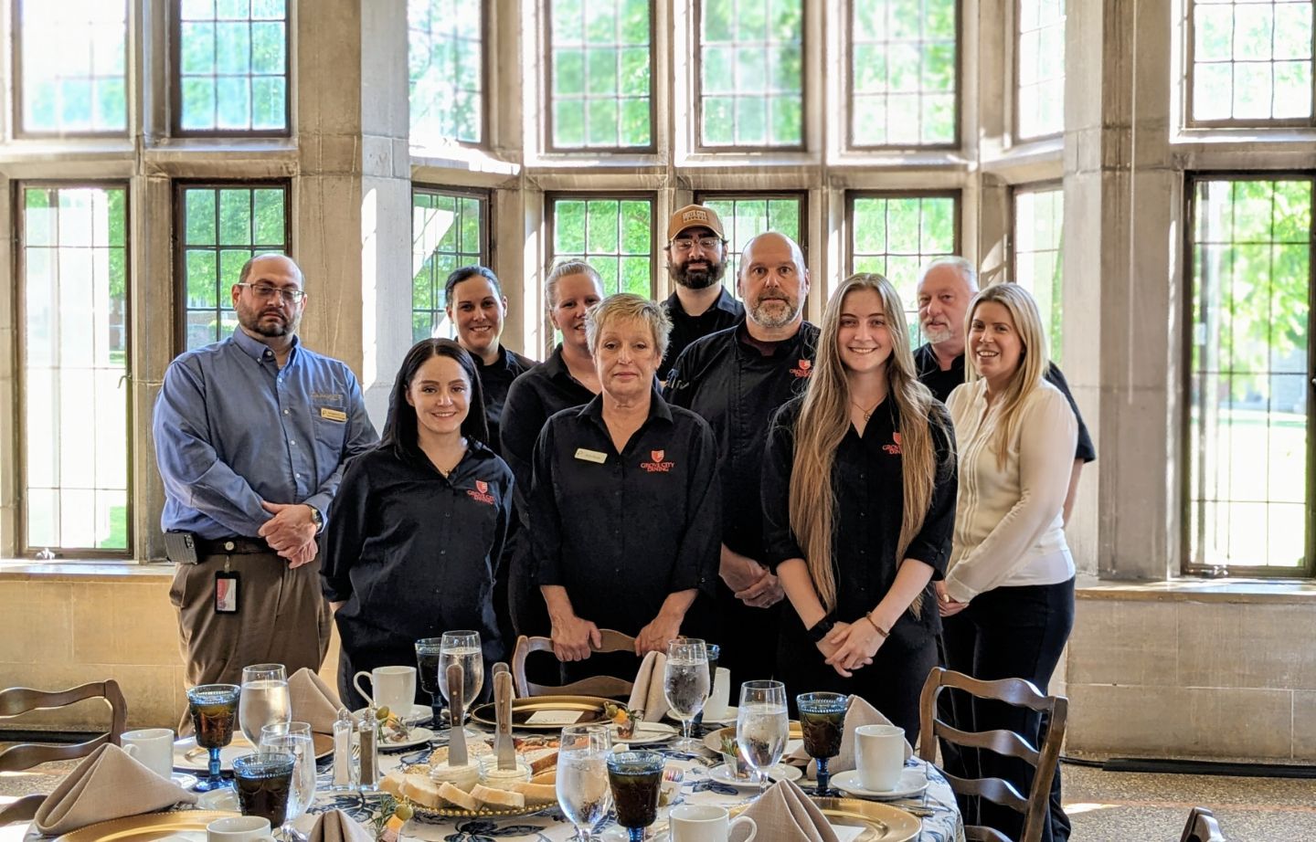 A group of diverse restaurant staff poses around a table set for a meal, with large windows in the background.