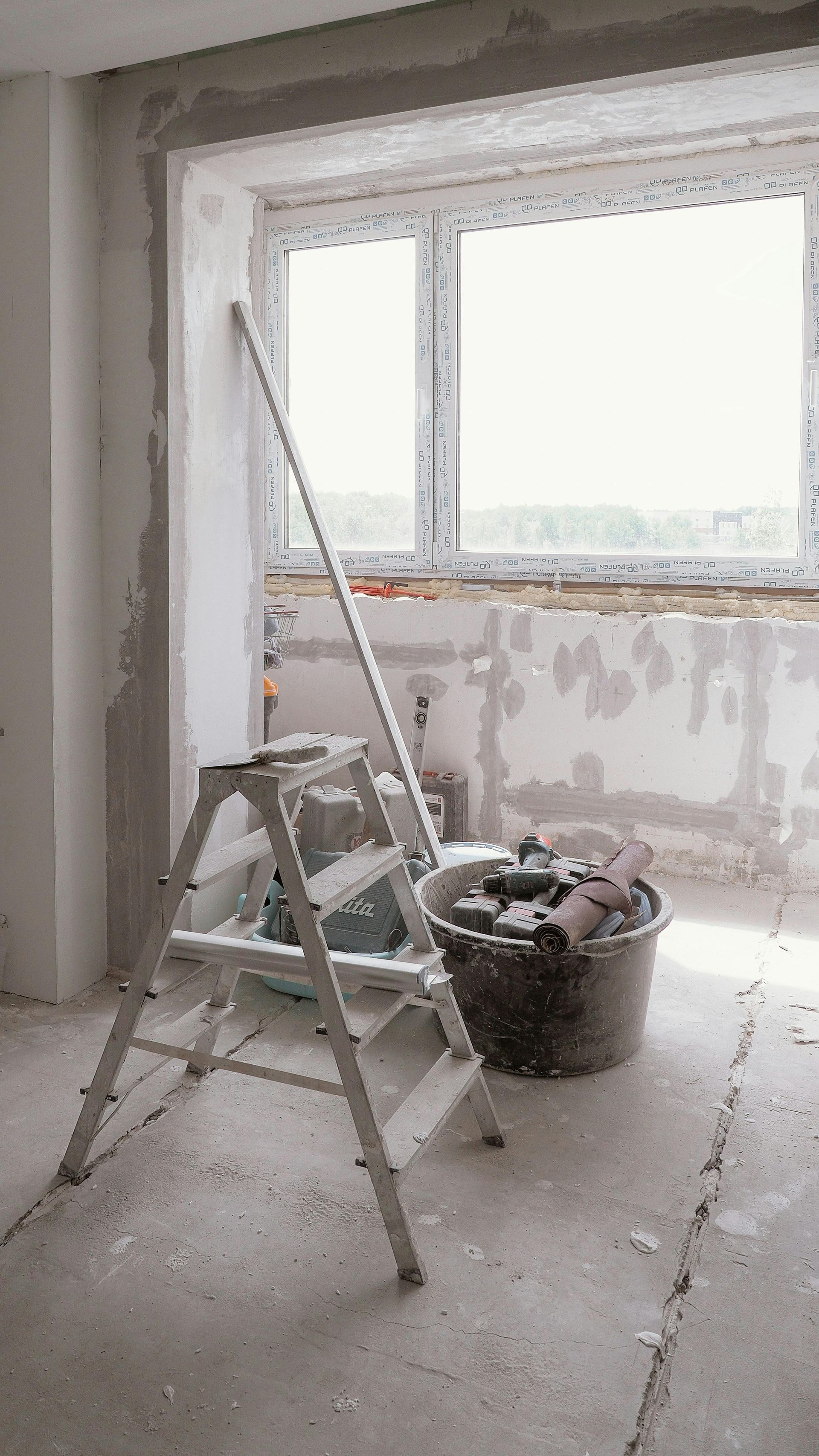 A renovation room with a ladder, bucket, and unfinished plaster walls by a window.