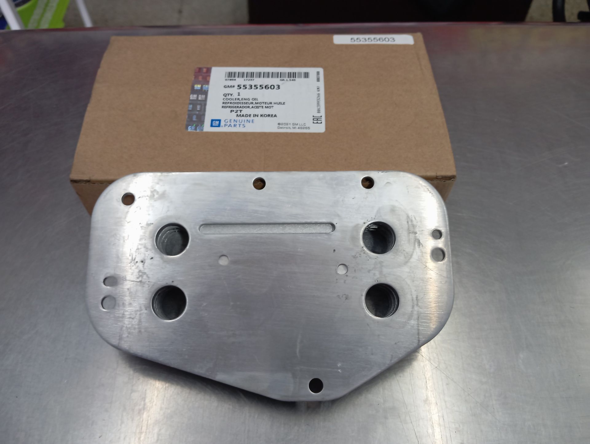 Pieza metálica con múltiples agujeros delante de una caja de cartón sobre una mesa.