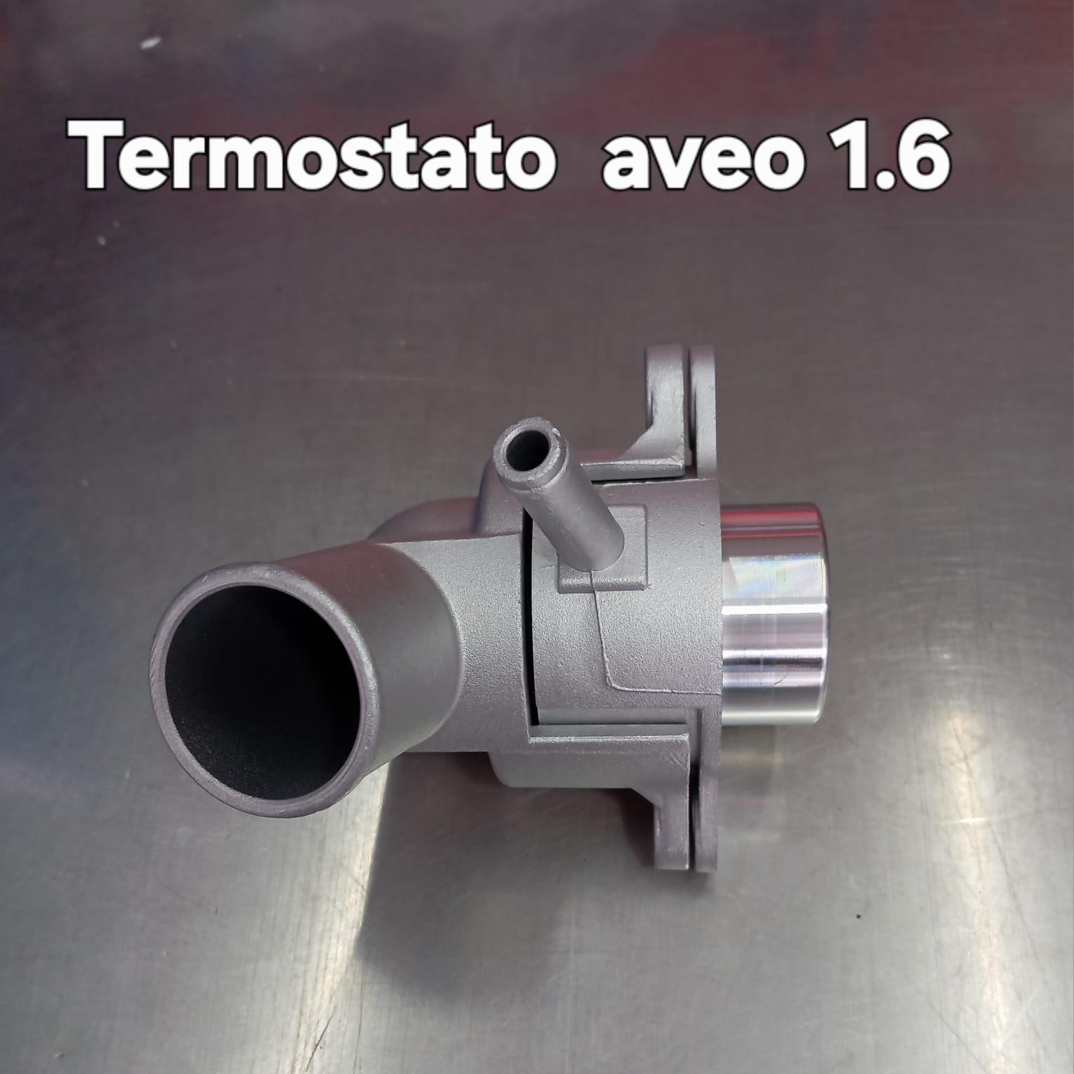 Termostato para motor Aveo 1.6. Pieza metálica plateada con varias conexiones de tubos.