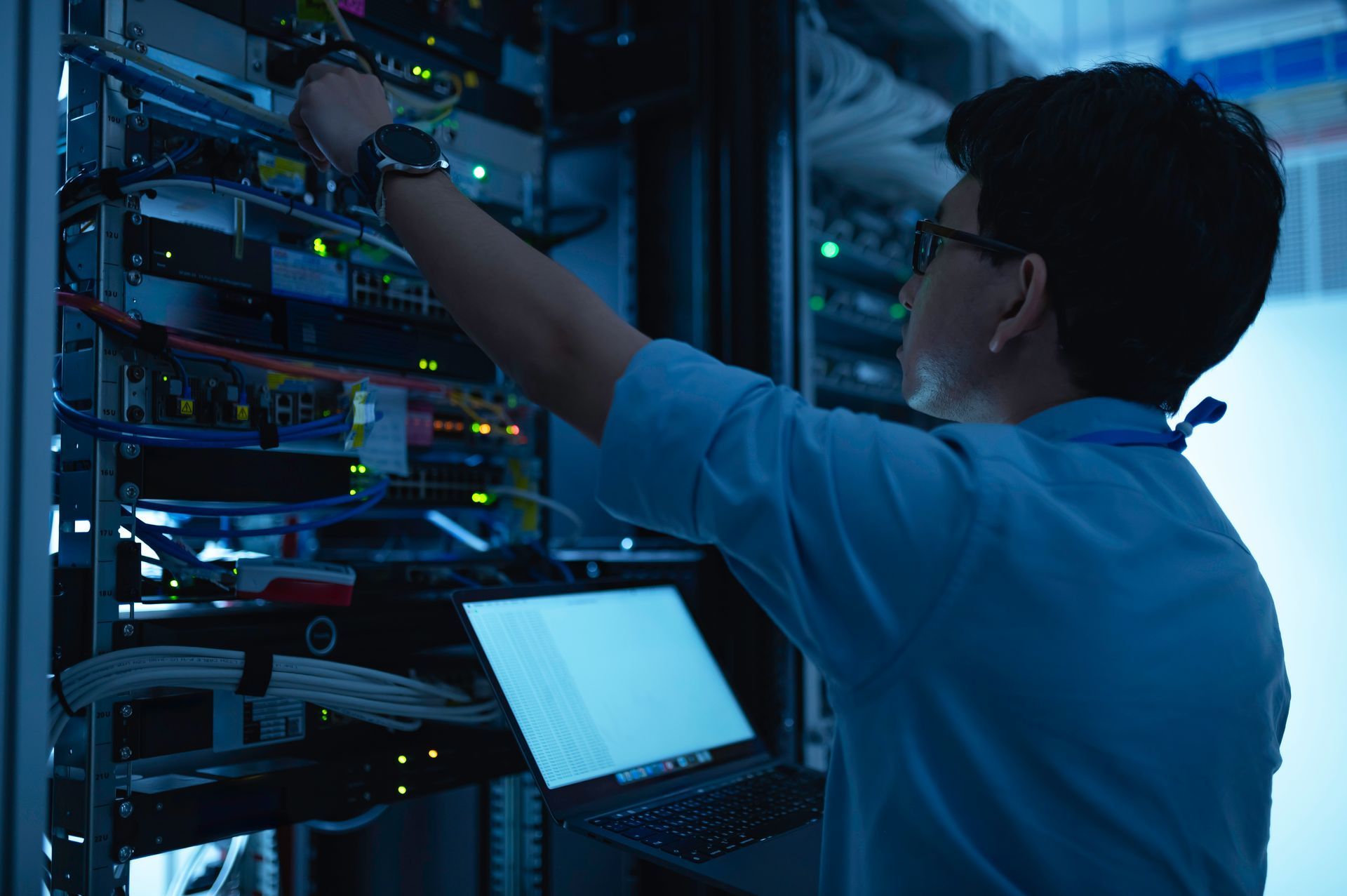 Technician working on server rack in a data center, holding a laptop and connecting cables.