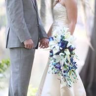 Wedding couple holding hands; bride in white gown with blue-and-white bouquet, groom in gray suit.