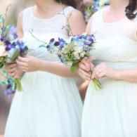 Two women in white dresses holding small purple and green bouquets outdoors