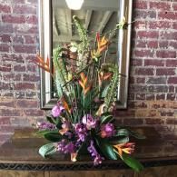 Colorful floral arrangement with orange, purple, and green flowers on a wooden table against a brick wall.