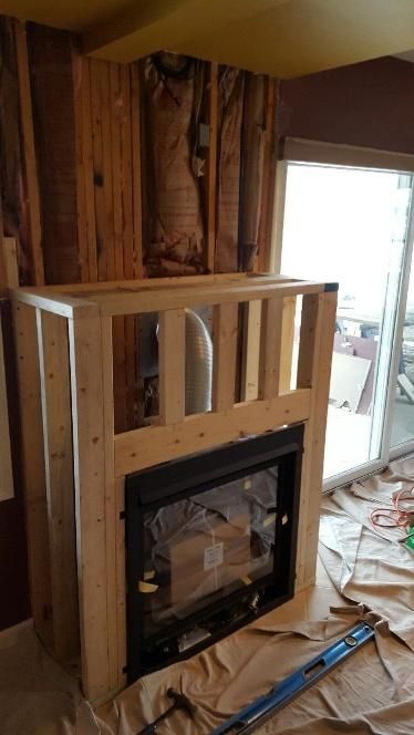 Wooden frame constructed around a black fireplace insert, with exposed wall behind and a sliding glass door to the right.
