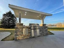 Outdoor kitchen with stone base, stainless steel grill, and white roof under a blue sky.