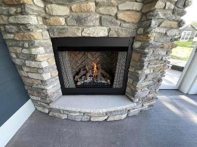 Stone fireplace with lit flames, set in a room with blue wall and gray flooring.