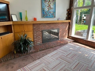 Living room with brick fireplace, built-in cabinets, colorful art, large window, and patterned carpet.