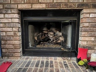 A brick fireplace with a black framed firebox, containing logs and a broken glass door.