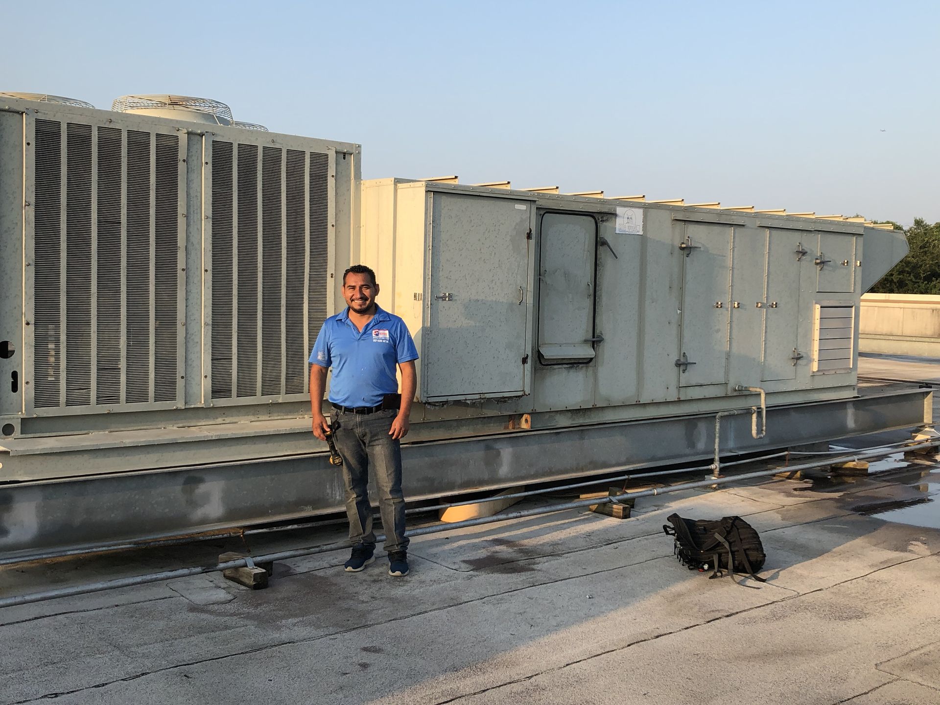 Man in blue shirt stands on a rooftop beside large HVAC unit.