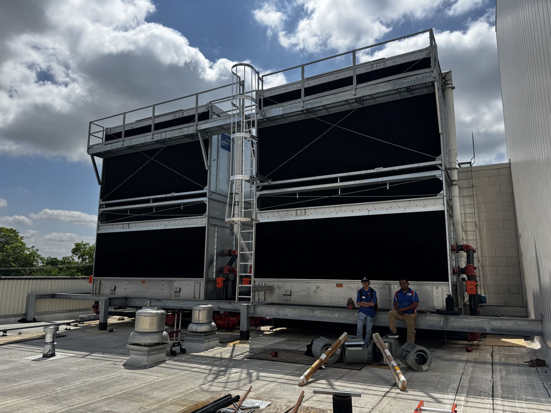 Two workers near a rooftop cooling tower, a large rectangular structure with black panels and metal framework.