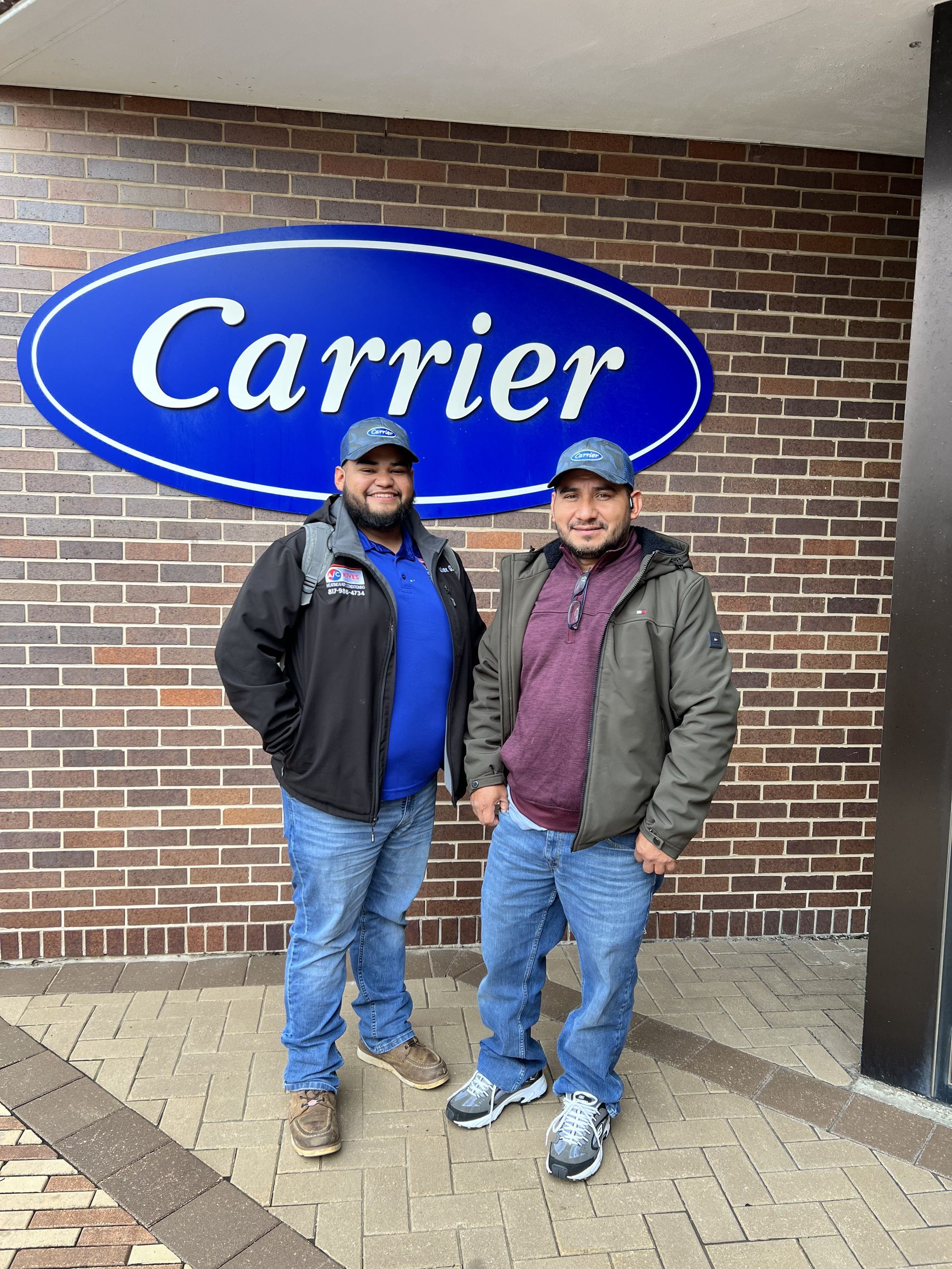 Two men in hats, standing in front of a Carrier sign on a brick building.