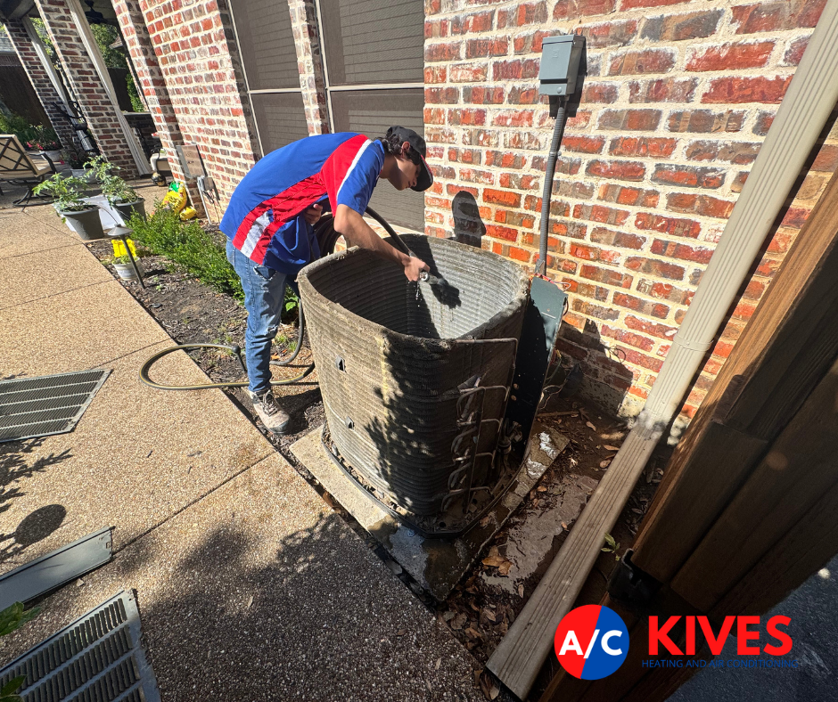 A man cleaning an outdoor AC unit with a brush, near a brick wall and garden.