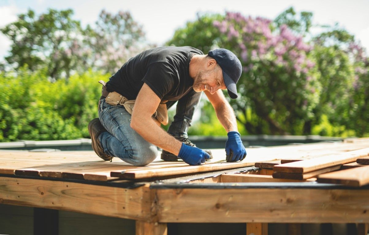 Man kneeling on a wooden deck, wearing gloves, cap, and tool belt, laying down wooden planks outdoors.