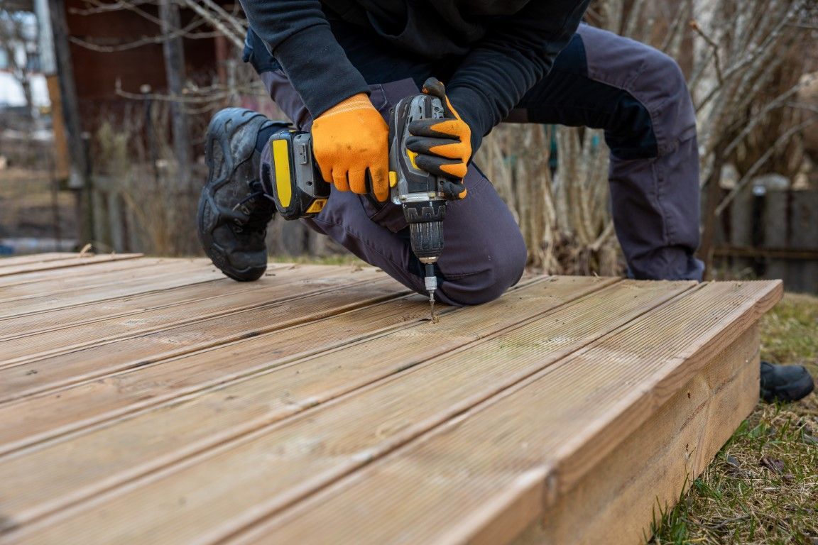 Person kneeling on a wooden deck, using a drill. Wearing gloves and dark work clothes. Outdoor setting.