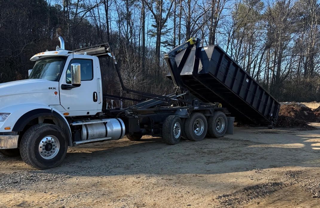 A roll off dump truck is parked in a gravel lot next to a dumpster.