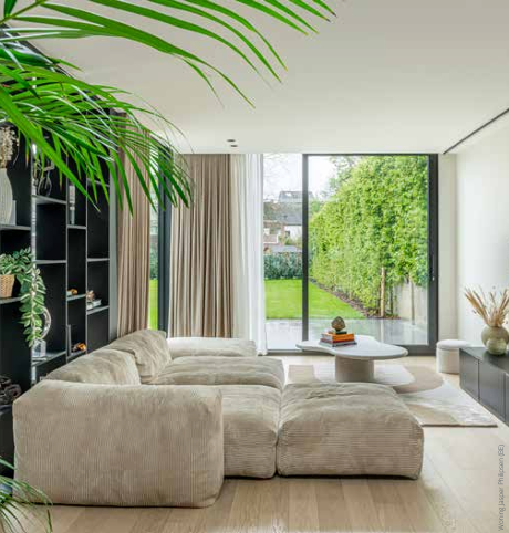 Moderne woonkamer met beige hoekbank, groot raam en uitzicht op de groene tuin.