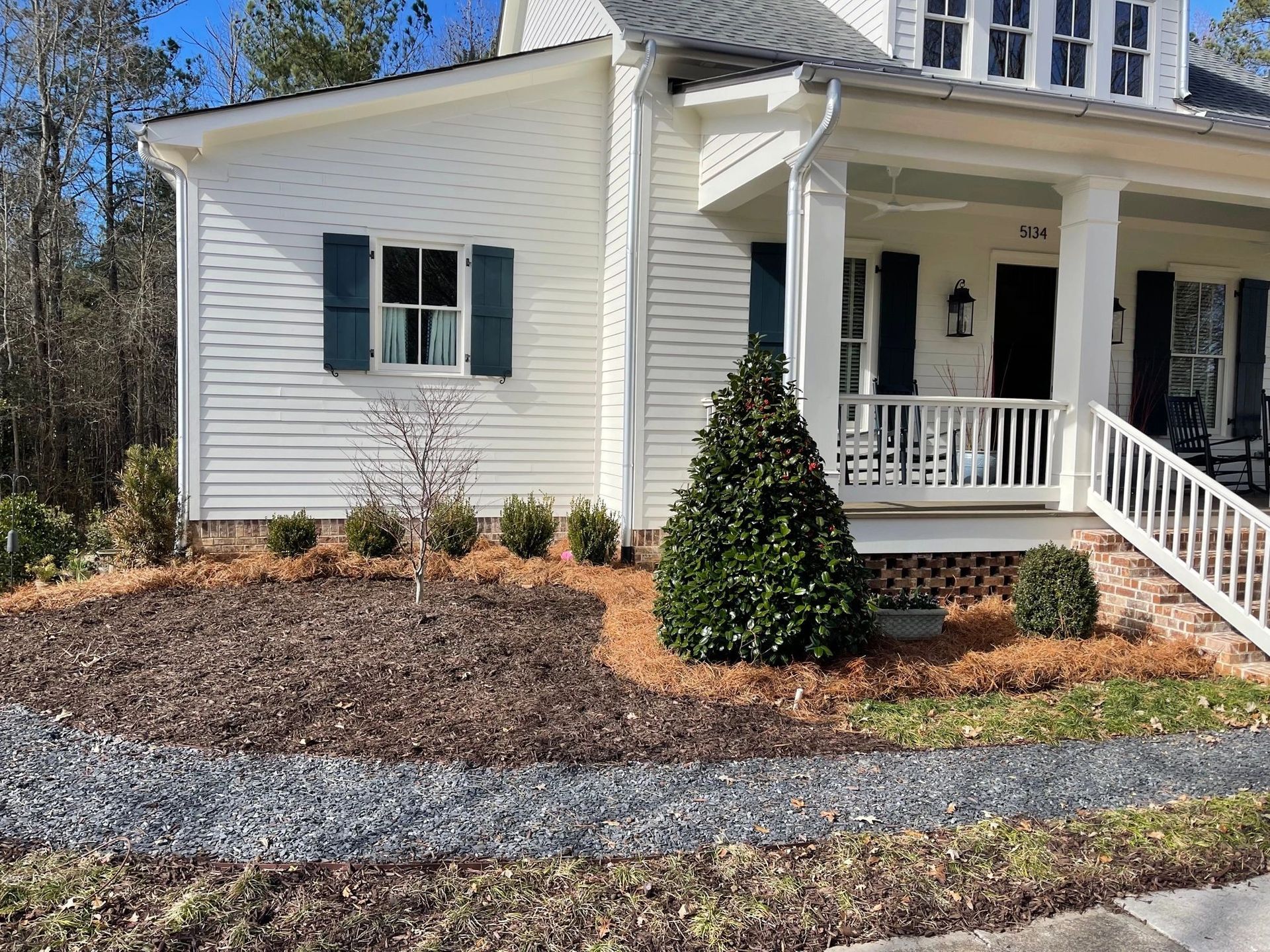A white house with dark shutters, a front porch, and a mulched garden bed in the front yard.