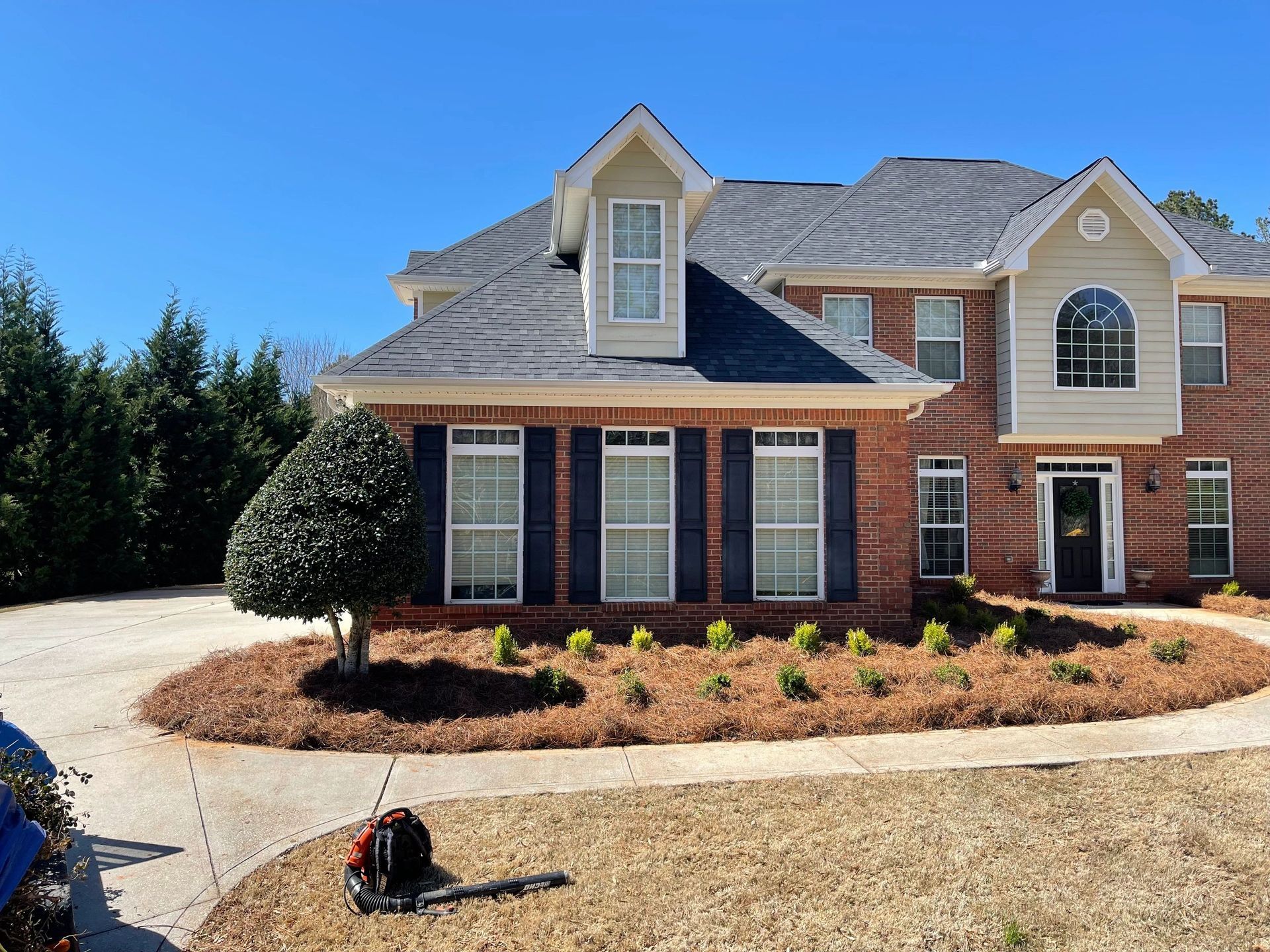 A two-story brick home with a landscaped front yard, a curved mulch bed, and a leaf blower resting on the lawn.