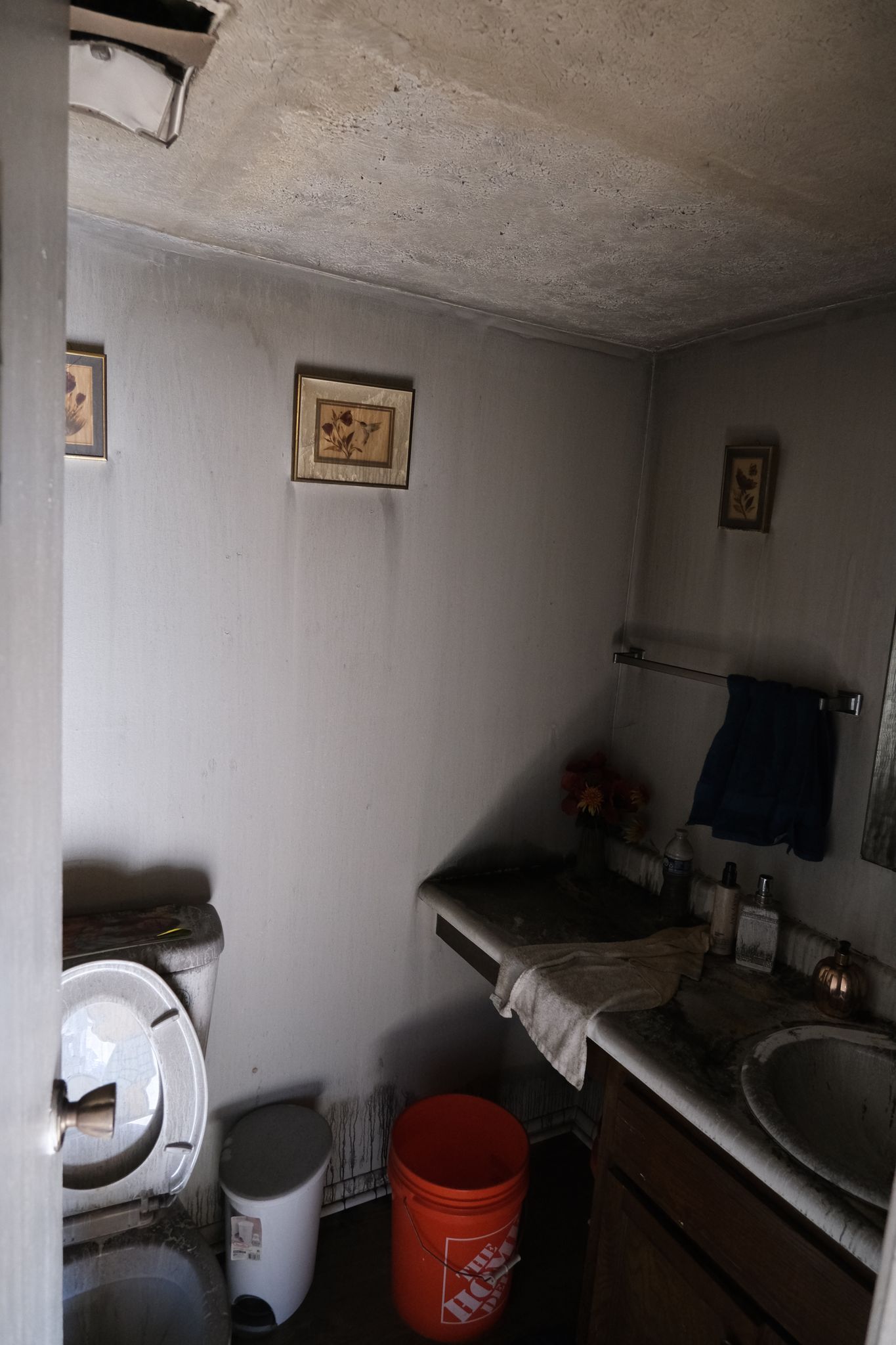 Dirty, small bathroom with a toilet, sink, and framed art on walls. Ceiling has a hole.