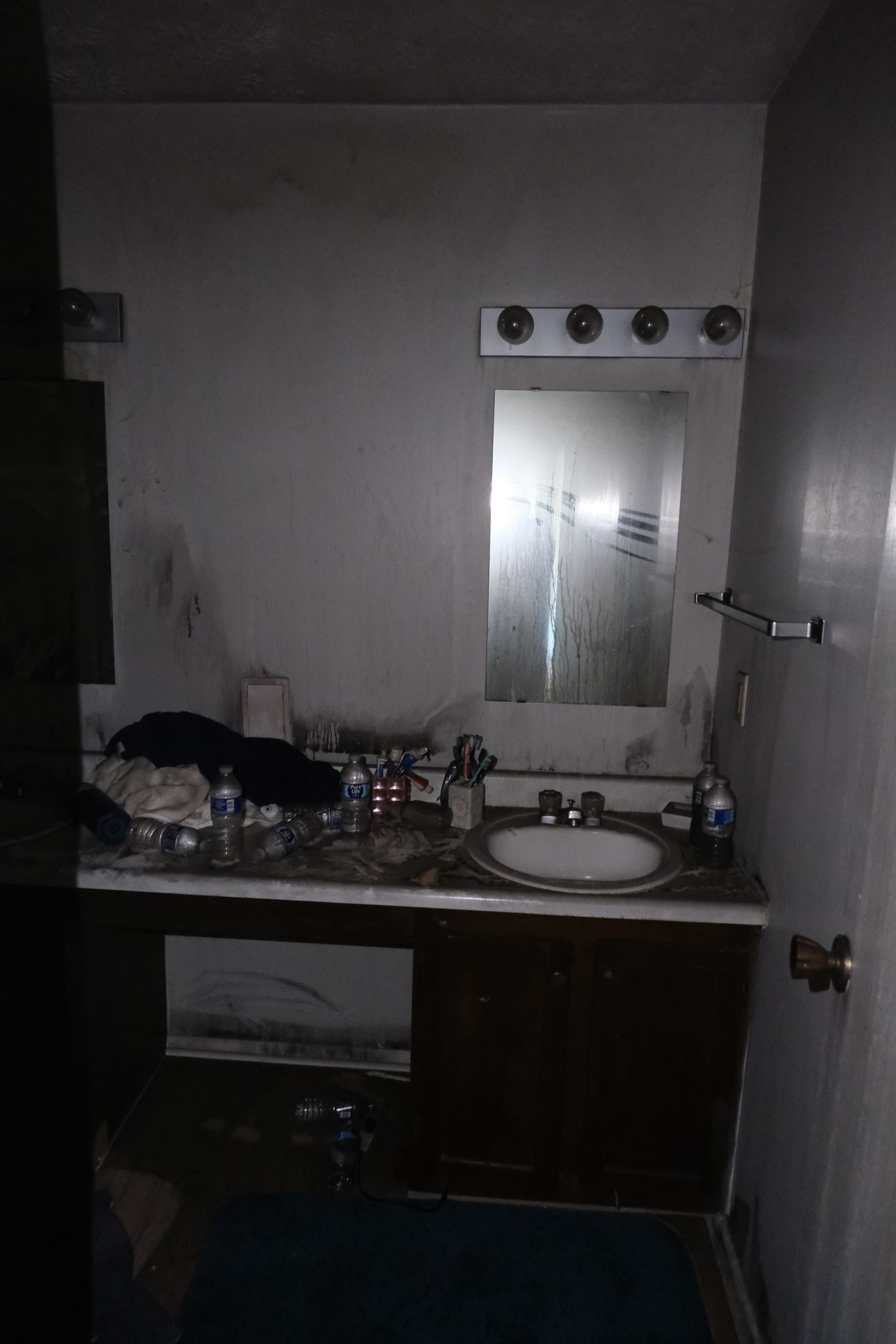 Bathroom: dirty walls, vanity with sink, mirror, towel rack, and cluttered countertop. Dark.