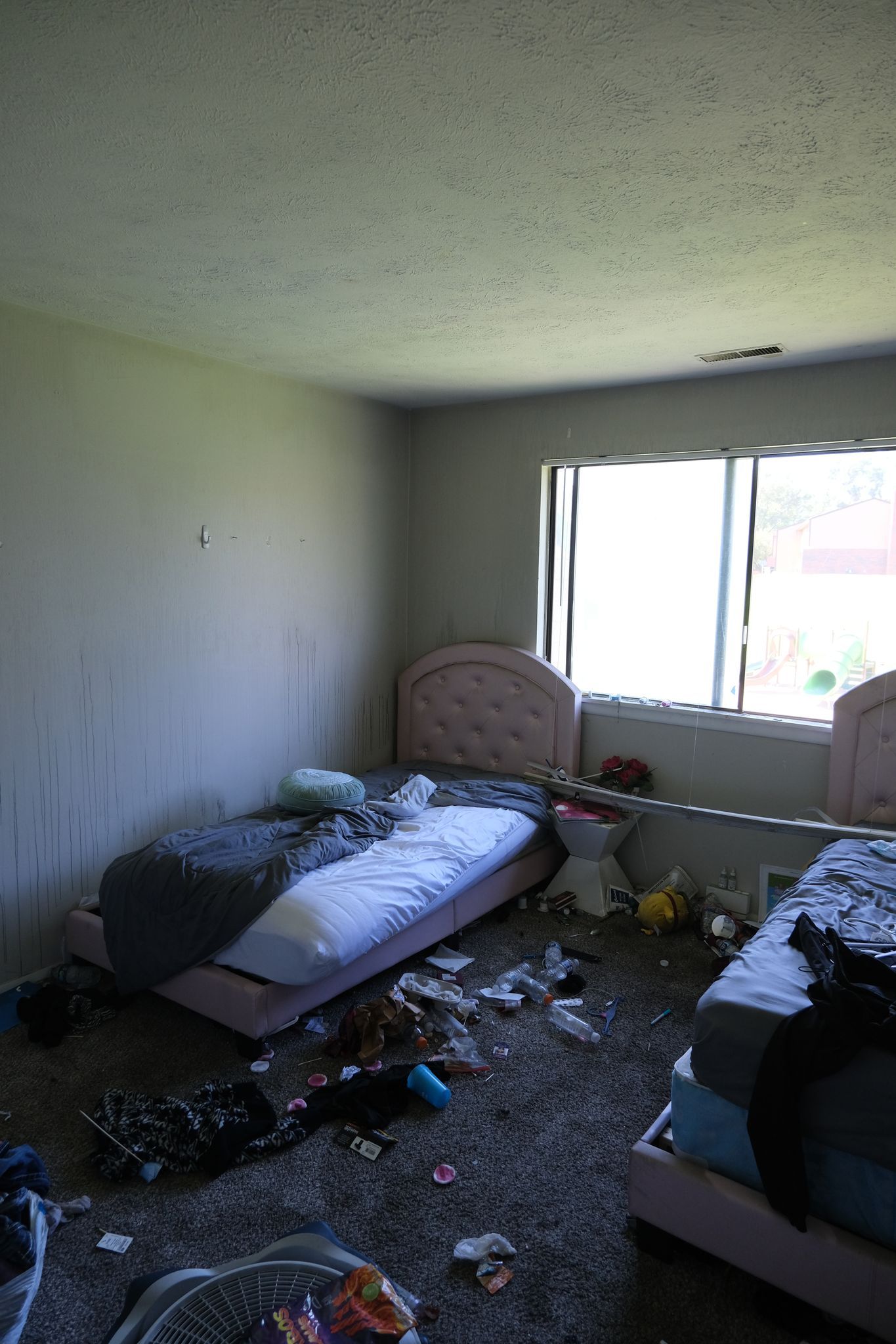 A messy bedroom with two beds, cluttered carpet, and a window letting in light.