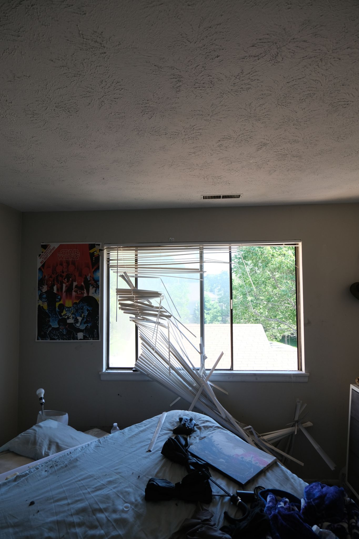 A messy bedroom with a broken window, debris on the bed, and colorful artwork.