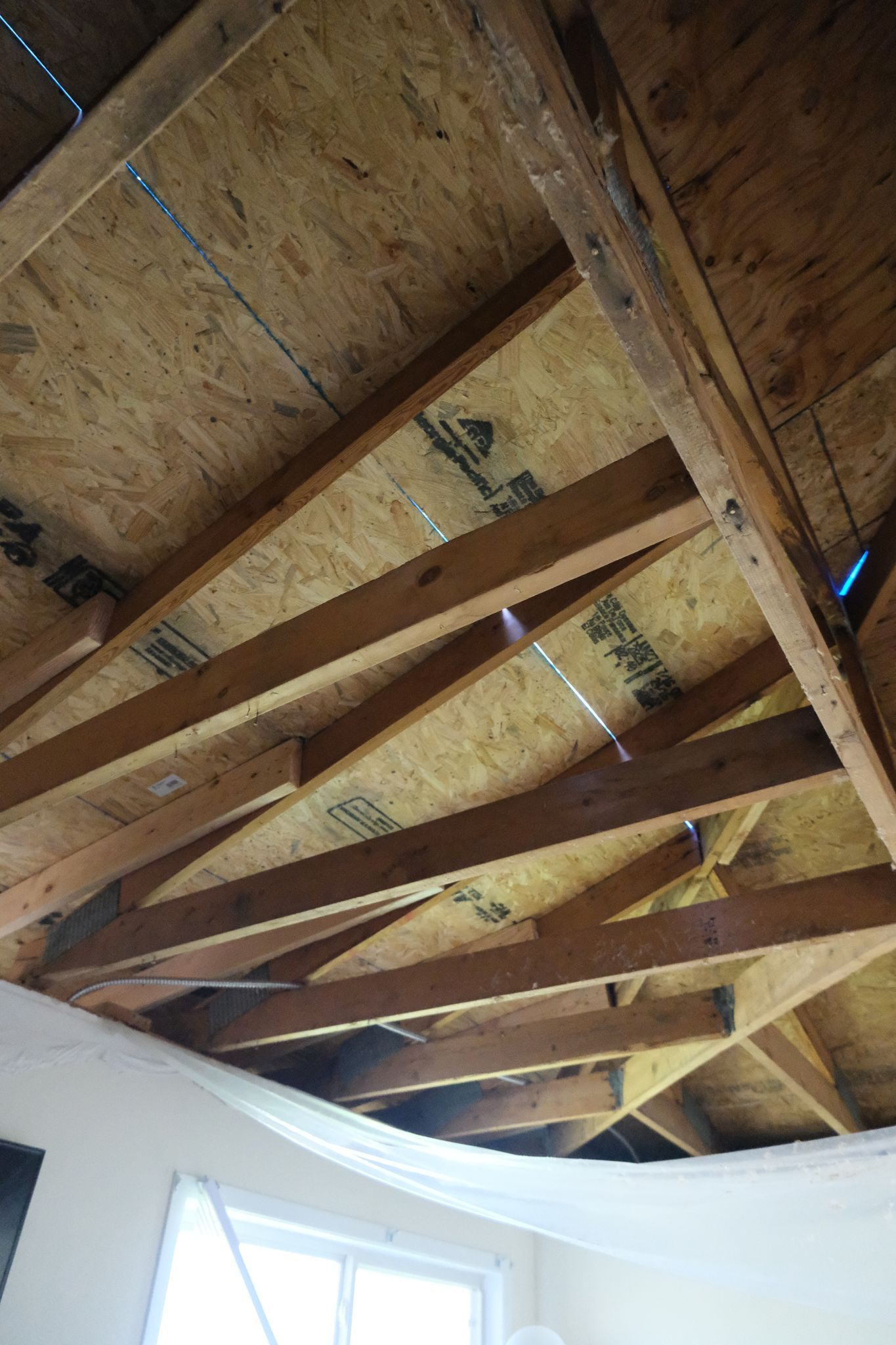 View from below of a wooden roof's structural beams and sheathing. Interior with natural light entering a window.