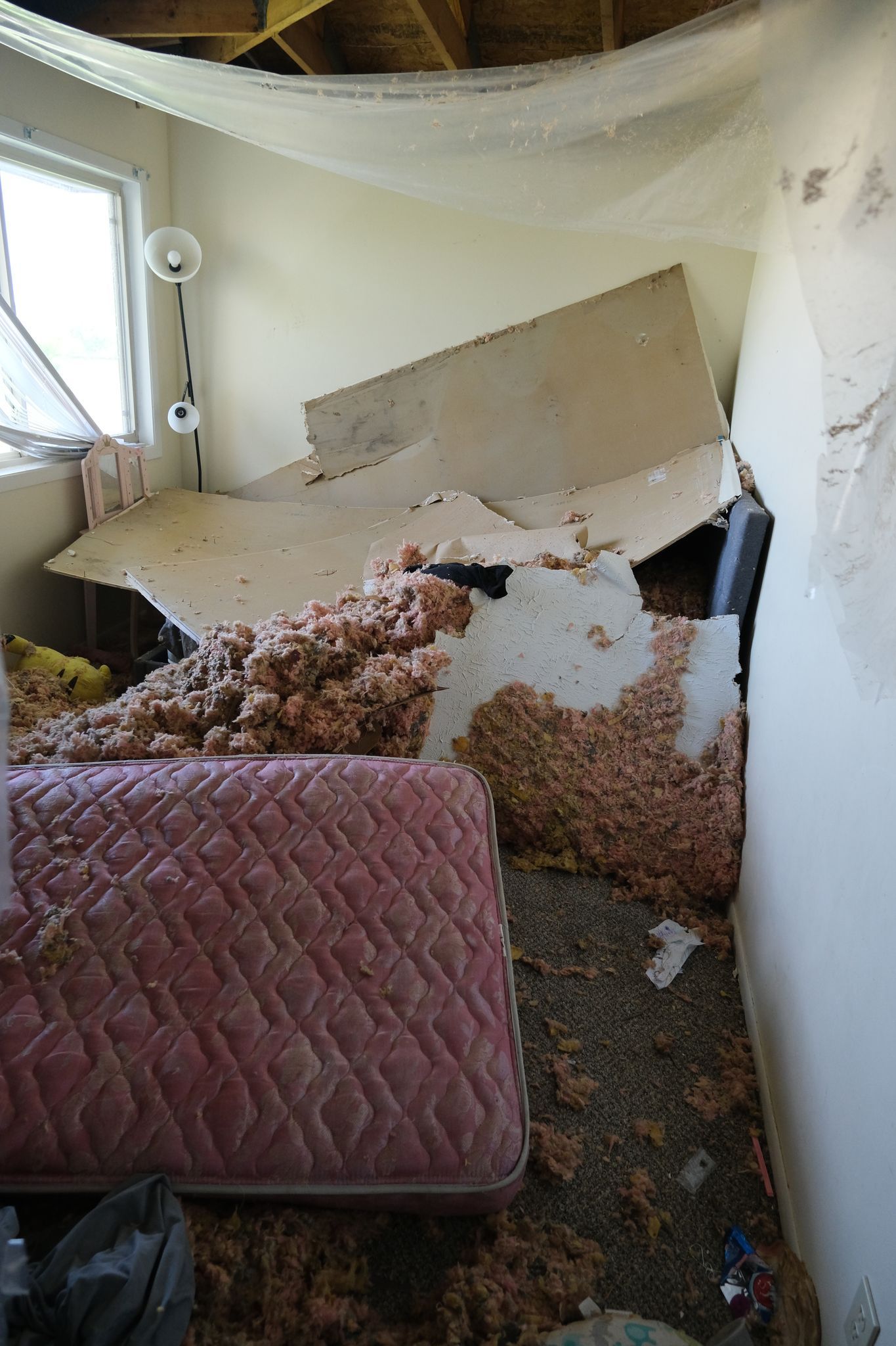 Room with collapsed ceiling and insulation debris on mattress, window in background.