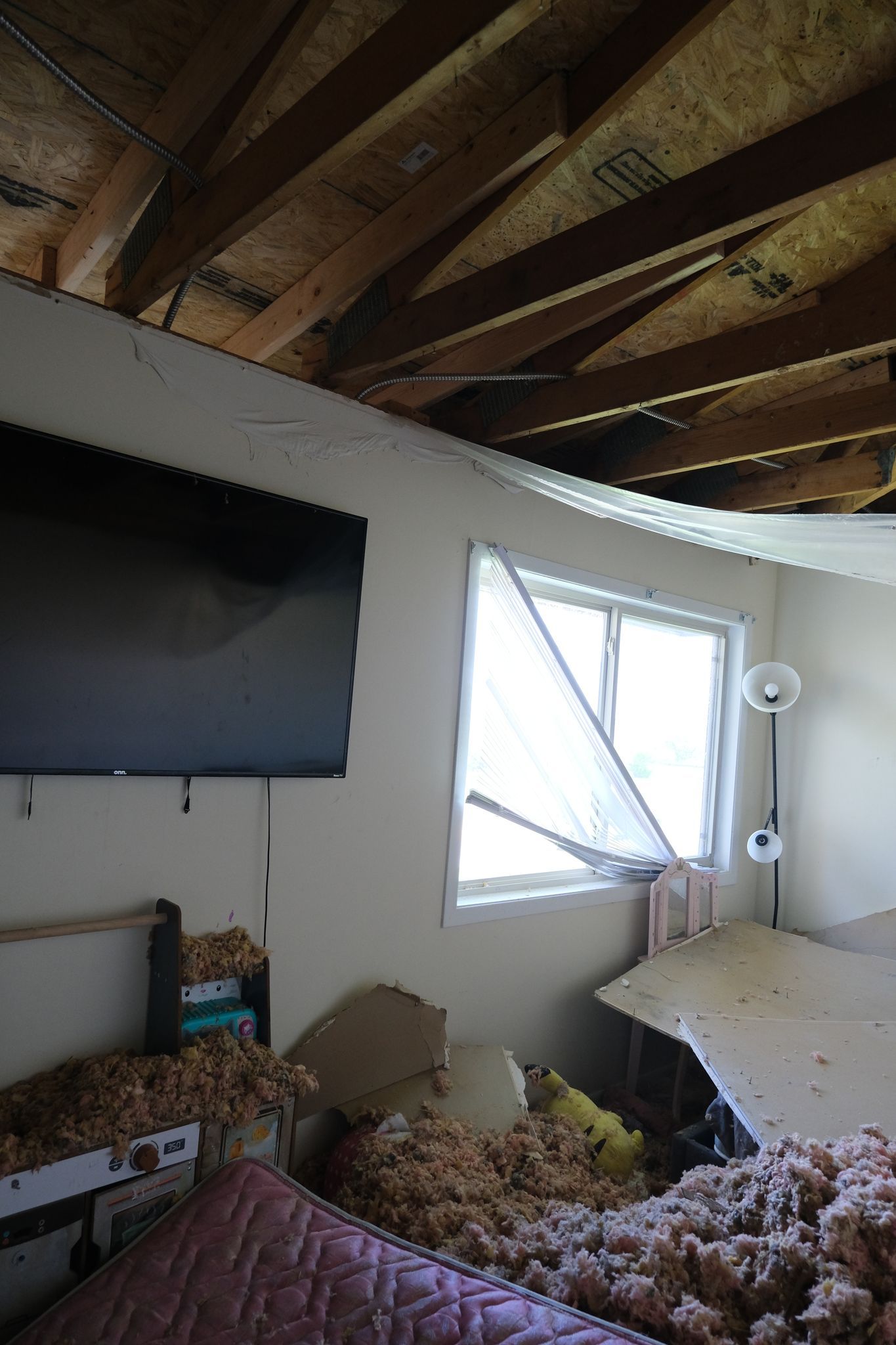 Room interior damaged, insulation and debris strewn, collapsed ceiling, window, and television on the wall.