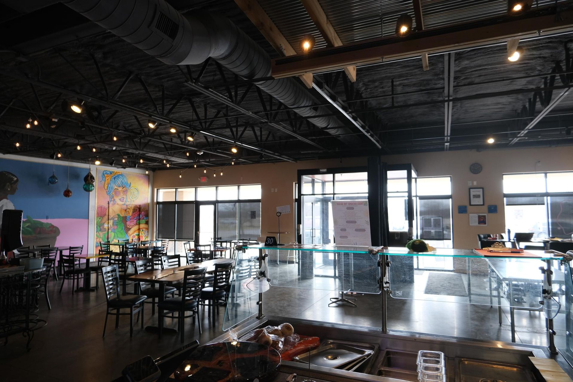 An open-concept restaurant with an industrial ceiling, dark walls, dining tables, and a mural on the far left wall.